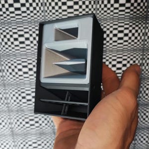 May include: A black and gray speaker enclosure with a unique design. The speaker has a rectangular shape with internal angled components. The background is a black and white checkered pattern.