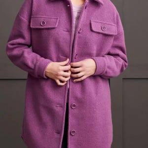 Könnte beinhalten: Eine lange, violette Jacke mit Knopfleiste, zwei Brusttaschen und einem Kragen. Die Jacke ist aus einem strukturierten Stoff gefertigt und wird über einem hellen Oberteil und einer schwarzen Hose getragen. Die Jacke reicht bis zur Mitte des Oberschenkels.