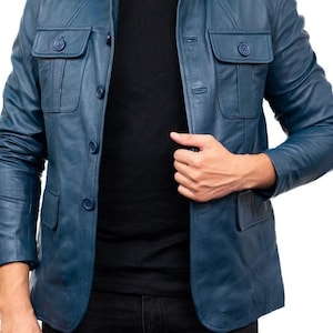 Puede incluir: Chaqueta de cuero azul con cierre de botones, dos bolsillos en el pecho y cuello. La chaqueta se lleva sobre una camiseta negra con cuello en V y pantalones negros. La chaqueta tiene un diseño clásico y a medida.