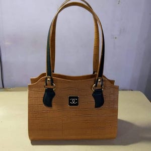 Könnte beinhalten: Eine hellbraune Handtasche mit texturiertem Krokodilleder-Muster, ausgestattet mit zwei hohen Tragegriffen mit schwarzen Akzenten an der Basis. Ein kleines, quadratisches schwarzes Emblem mit silberfarbenem Logo ist mittig auf der Vorderseite angebracht.