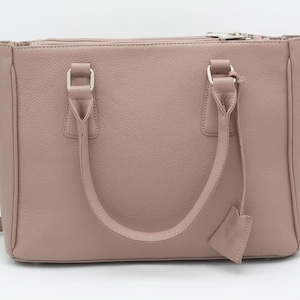 May include: A dusky pink handbag with a textured finish. The bag has a top zip closure, two handles, and a detachable shoulder strap. Silver-tone hardware accents the bag's design.