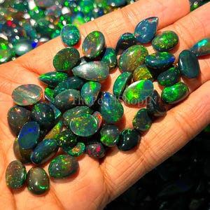 May include: A collection of polished black opal gemstones, displaying flashes of green, blue, and red. The opals are oval and teardrop-shaped, in various sizes and patterns. The gemstones are held in a person's hand.