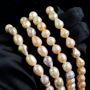 Puede incluir: Cuatro hilos de perlas en tonos crema, melocotón y lavanda. Las perlas son de formas irregulares y están ensartadas. El fondo es negro. Las perlas se miden en cm.