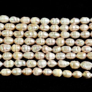 Puede incluir: Primer plano de hileras de perlas de agua dulce ovaladas, de color blanquecino con un ligero tinte rosado. Las perlas están ensartadas, listas para la creación de joyas. Las perlas tienen una superficie lisa y ligeramente texturizada. Fondo negro.