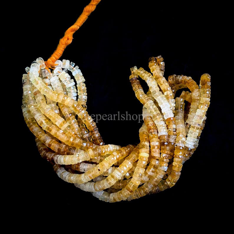 May include: A collection of faceted citrine gemstone beads in shades of yellow and brown, strung together with an orange cord. The beads are small and square-shaped, with a polished finish. The background is black.