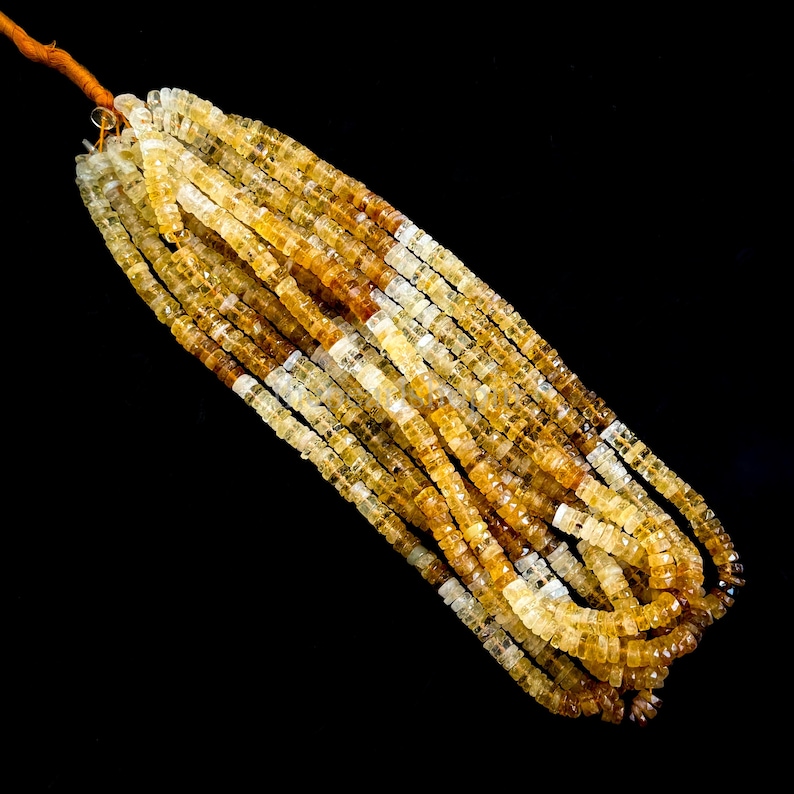 May include: Strands of faceted citrine gemstone beads in shades of yellow and brown. The beads are square-shaped and strung together, creating a textured and colorful display. The gemstones are displayed against a black background.