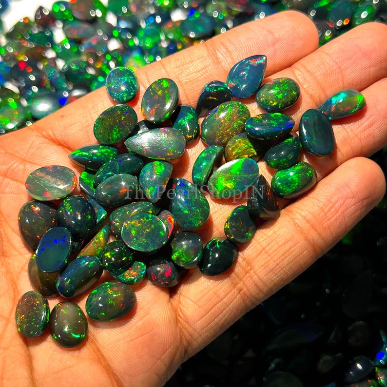 May include: A collection of polished black opal gemstones, displaying flashes of green, blue, and red. The opals are oval and teardrop-shaped, in various sizes and patterns. The gemstones are held in a person's hand.