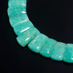 May include: A section of a necklace made of polished, rectangular amazonite beads. The beads are a vibrant turquoise-green color with subtle variations in tone. The necklace is displayed against a black background, highlighting the color and texture of the gemstones.