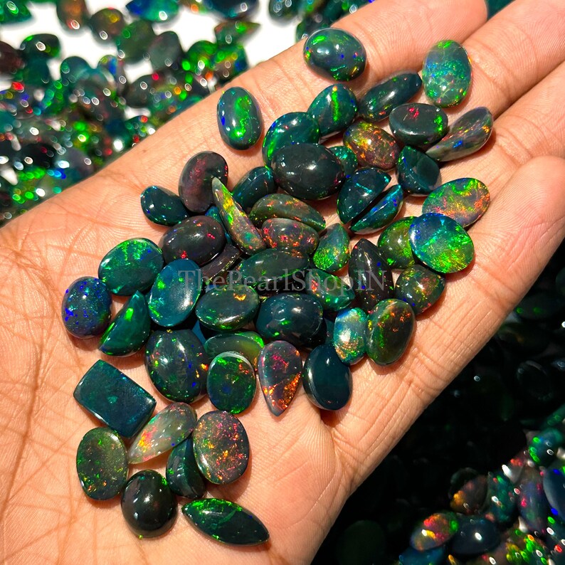 May include: A selection of black opal gemstones, displaying a range of shapes and sizes, with striking flashes of colour. The opals, in shades of green, blue, and red, are held in a hand, highlighting their natural iridescence and unique patterns.