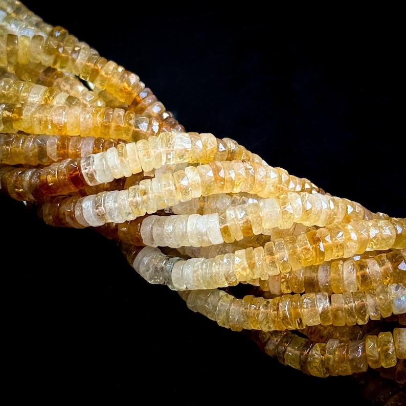 May include: Close-up of strands of citrine gemstone beads. The beads are a mix of light yellow, amber, and clear tones. Each bead is a small, faceted, disc shape. The background is solid black, highlighting the color and texture of the gemstones.
