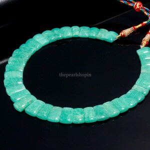 May include: A necklace featuring rectangular, light green amazonite beads. The beads are strung together, forming a circular shape. The necklace is displayed on a black surface. The string has red, blue, and gold accents.