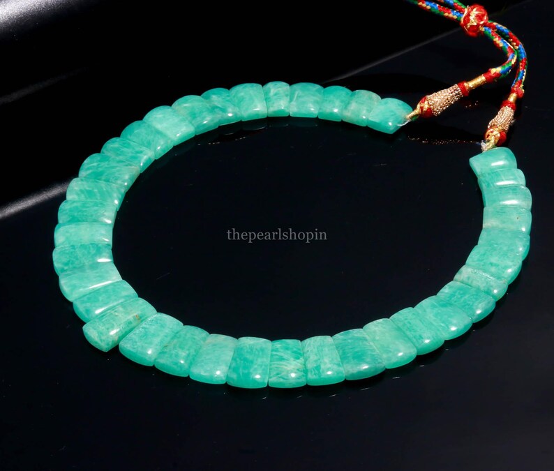 May include: A necklace featuring rectangular, light green amazonite beads. The beads are strung together, forming a circular shape. The necklace is displayed on a black surface. The string has red, blue, and gold accents.