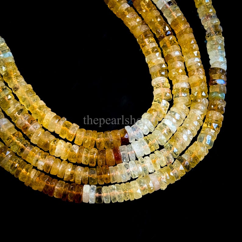 May include: Close-up of several strands of faceted, golden-yellow gemstone beads. The beads are strung together, creating a layered effect. The color varies from light yellow to a deeper amber hue, with some beads appearing translucent. The background is black.