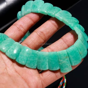 May include: A light green Amazonite bracelet. The bracelet is composed of rectangular, polished stones with a scalloped edge design. The stones are a light, opaque green color. The bracelet is held in a hand against a dark background.