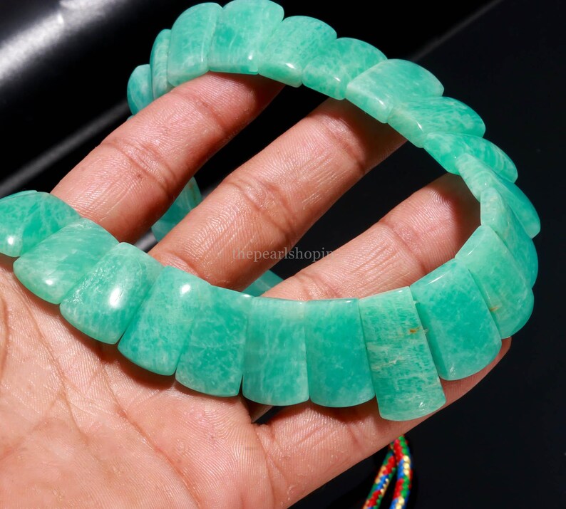 May include: A light green Amazonite bracelet. The bracelet is composed of rectangular, polished stones with a scalloped edge design. The stones are a light, opaque green color. The bracelet is held in a hand against a dark background.