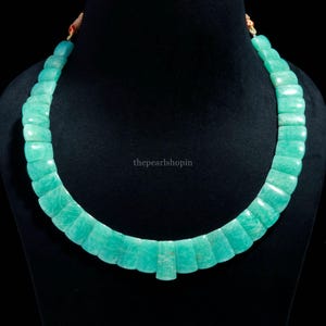 May include: A turquoise-colored necklace composed of rectangular, polished stones. The stones are strung together to form a curved shape. The necklace is displayed on a black mannequin. The text "thepearlshopin" is visible on the image.