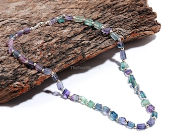 May include: A necklace featuring rectangular fluorite beads in shades of purple, green, and blue, strung together with small silver-colored beads. The necklace is displayed on a piece of textured wood.