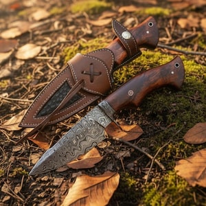 May include: A handcrafted hunting knife with a patterned blade and a dark wooden handle. The knife is accompanied by a brown leather sheath with a cross design. The knife and sheath are set on a bed of leaves and moss.