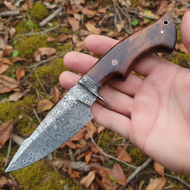 May include: A handcrafted knife with a patterned steel blade and a dark wood handle. The blade has a unique swirling design. The handle is a rich brown color, and the knife appears to be held in a hand.