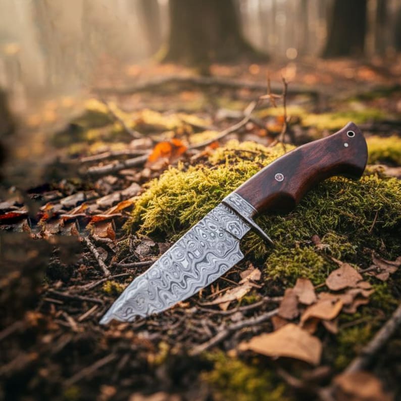 May include: A hunting knife with a patterned steel blade and a dark wood handle rests on a bed of green moss and fallen leaves. The knife has a curved blade and a bolster where the blade meets the handle. The background is a blurred forest scene.
