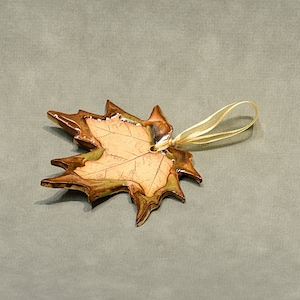 May include: A maple leaf ornament with a gold ribbon for hanging. The leaf has a detailed design with brown, green, and tan colours. The ornament is likely made of ceramic or a similar material.