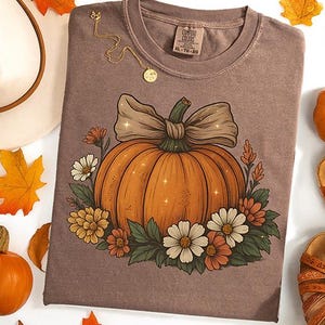 Puede incluir: Una camiseta marrón con un diseño de calabaza adornada con un lazo y flores. La imagen incluye hojas de otoño, un sombrero, sandalias y un collar dorado. La camiseta tiene el texto "COMFORT COLORS" e información de tallas.