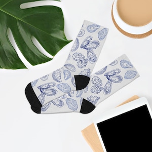 May include: A pair of white socks with a blue oyster shell pattern. The socks have black heels and toes. The socks are laying on a white surface with a green leaf, a cup of coffee, and a tablet in the background.