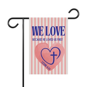 May include: A garden flag with a pink and white striped background. The flag has the words "WE LOVE" in blue, with the text "BECAUSE HE LOVED US FIRST" below. A pink heart with a blue cross is in the center, with the text "1 JOHN 4:19" below.