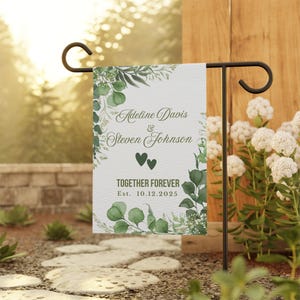 May include: A white garden flag with green foliage and the names "Adeline Davis & Steven Johnson" printed in elegant script. The flag also features two green hearts and the words "TOGETHER FOREVER" with the date "Est. 10.12.2025".
