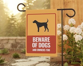 Puede incluir: Una bandera de jardín rectangular con la silueta negra de un perro sobre un fondo amarillo, encima de un rectángulo rojo con el texto "BEWARE OF DOGS AND OWNERS TOO". La bandera está sujeta a una estaca de jardín de metal negro.