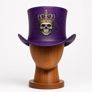 Op de afbeelding: Een paarse hoge hoed met een centrale afbeelding van een menselijke schedel die een gouden kroon met paarse accenten draagt. De schedel is omringd door donkere, gedetailleerde filigraanpatronen. De hoed heeft een gestructureerde bol en een gebogen rand.