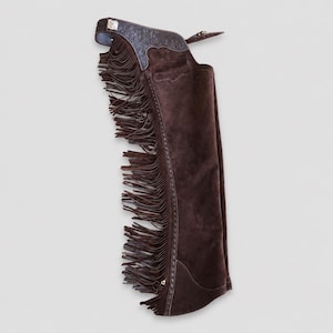 May include: A dark brown suede leather chap featuring long fringe along the outer leg. The upper section displays intricate floral tooling on the leather yoke, complemented by a silver-toned buckle and a reinforced stitched seam along the side.
