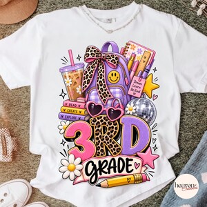 May include: A white t-shirt featuring a colorful graphic of a purple backpack with a leopard print bow, a coffee cup, heart-shaped sunglasses, a disco ball, and school supplies. The text reads 3RD GRADE and Dream Learn Be Kind.