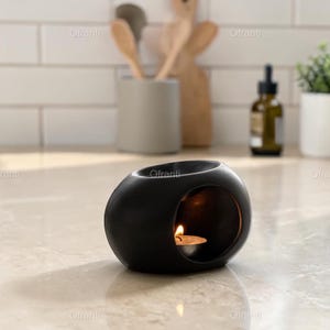May include: A black, oval-shaped essential oil diffuser with a small candle lit inside. The diffuser has a smooth, matte finish and a circular opening. The background includes wooden spoons, a small plant, and a bottle.