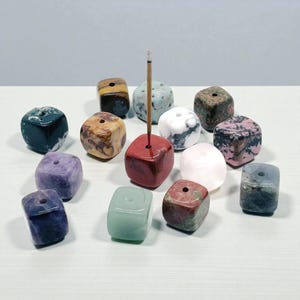 May include: A selection of cube-shaped incense holders in diverse colours and patterns. The holders are crafted from stone and have a hole for incense sticks. A red holder holds a lit incense stick. The holders are displayed on a white surface.