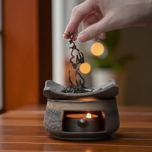 Puede incluir: Un difusor de aceites esenciales de cerámica con una vela de té encendida en su interior. El difusor es de color gris oscuro y marrón con una superficie texturizada. Una mano coloca hierbas secas encima.