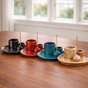 Puede incluir: Colección de portainciensos de cerámica en varios colores, incluyendo negro, rojo, verde azulado y beige. Cada soporte tiene un plato a juego con un patrón decorativo y un pequeño palito de incienso. Dispuestos sobre una superficie de madera.
