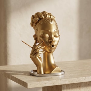 May include: A gold-colored sculpture of a head and hand, featuring a stylized face with closed eyes and pursed lips. The hand is raised near the face, holding a thin, straight stick. The figure is mounted on a circular, silver-toned base.