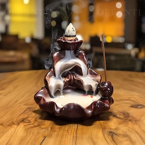 Könnte beinhalten: Ein brauner Keramik-Räuchergefäß mit einem Wasserfall-Design. Weißer Rauch steigt aus dem Brenner auf, mit einem Räucherkegel an der Spitze. Ein angezündeter Räucherstäbchen ist an der Seite platziert. Der Brenner steht auf einer Holzoberfläche.