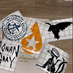 May include: A collection of vinyl decals featuring various designs including a circular Wishbone Sailing Club logo, an orange teardrop with a white star, a black silhouette of a creature, the text Conan Gray, and a black silhouette of a figure in a hat.