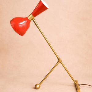 May include: A red and gold desk lamp with a cone-shaped shade and a curved, adjustable arm. The lamp has a brass-colored base and accents. The shade is a glossy red, and the interior is white.