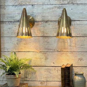 May include: Two brass cone-shaped wall sconces with a perforated design, mounted on a weathered wood wall. The sconces are lit, casting a warm glow. A potted fern, books, and a vase are on a wooden surface.