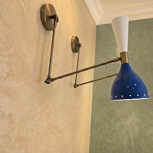 Peut inclure: Une applique murale dotée d'une structure en laiton à deux bras avec des articulations réglables. Le luminaire présente une partie supérieure conique blanche et un abat-jour bleu en forme de cloche avec des perforations circulaires.