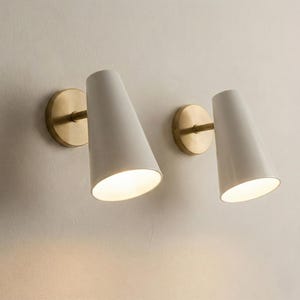May include: Two white, cone-shaped wall sconces with brass-coloured circular backplates and arms. The sconces are mounted on a light-coloured wall, and the lights are illuminated.