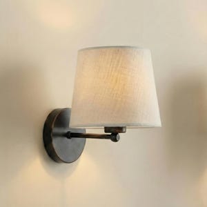 May include: A wall-mounted light fitting featuring a beige fabric lampshade. The fixture has a dark bronze finish, including the circular base and the arm supporting the shade. The lampshade is a classic tapered shape.