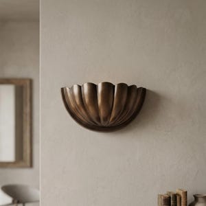 May include: A bronze-colored wall sconce with a scalloped, shell-like design. The fixture is mounted on a neutral-toned wall, showcasing its decorative, organic form. The sconce is a stylish addition to any interior space.