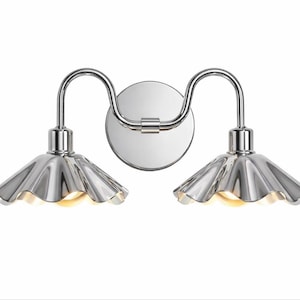 May include: A polished chrome wall light fitting featuring a circular mounting plate and two curved arms extending to support two downward-facing, fluted, bell-shaped shades with a reflective metallic finish and warm internal illumination.