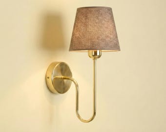 May include: A wall-mounted sconce with a gold-toned metal finish. It features a curved arm supporting a lampshade with a brown fabric cover. The sconce has a circular base and a simple, elegant design, suitable for interior lighting.