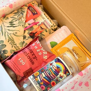 May include: A gift box containing a glass tumbler with a book and rainbow graphic labelled THE BOOK LOVER, a pink bag of Popcorn Shed Berry-licious popcorn, a London Fruit & Herb tea bag, an Aliver Crystal Collagen Gold mask, and a floral envelope.
