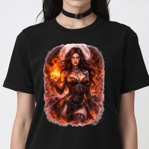 Puede incluir: Camiseta negra con una ilustración de fantasía de una mujer con alas de fuego y una llama. La obra de arte está ambientada sobre un fondo de llamas. La mujer lleva un corsé oscuro y tiene el pelo largo y rojo.